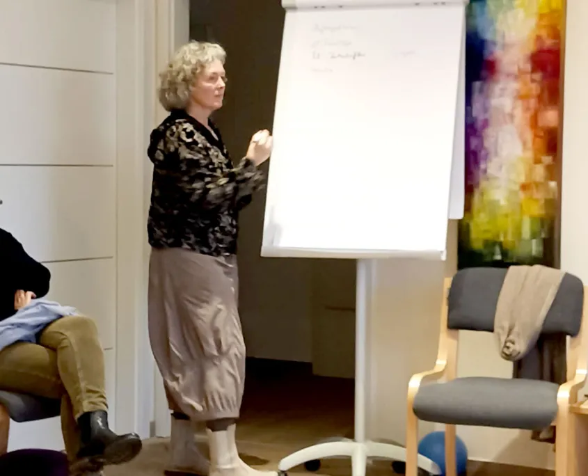 Hebamme Trauma-Fachberaterin - Coaching, Beratung, Kurse und Lesungen - Eine Frau mit kurzen, lockigen grauen Haaren steht vor einer Tafel und schreibt darauf, während sie eine kleine Gruppe anspricht. Sie trägt eine dunkel gemusterte Jacke und einen langen, hautfarbenen Rock. Rechts hängt hinter einem leeren Stuhl ein farbenfrohes abstraktes Gemälde an der Wand.