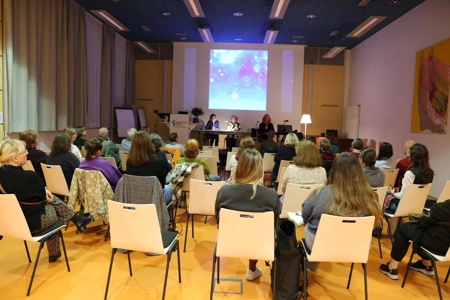 Hebamme Trauma-Fachberaterin - Coaching, Beratung, Kurse und Lesungen - Eine Innenraumaufnahme zeigt eine Präsentation oder Vorlesung in einem modernen Hörsaal mit blauer Decke und gelbem Boden. Das Publikum, von hinten gefilmt, sitzt in Reihen weißer Stühle vor einer Bühne, auf der drei Redner hinter einem langen Tisch Platz genommen haben. Im Hintergrund wird ein farbenfrohes kosmisches Bild auf eine große Leinwand projiziert.