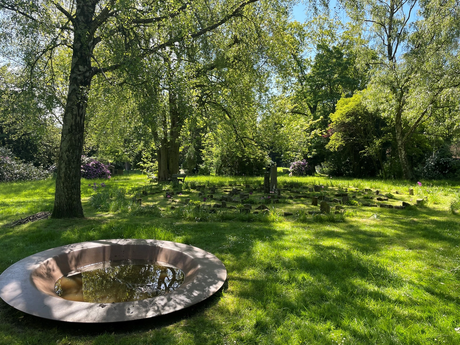 Hebamme Trauma-Fachberaterin - Coaching, Beratung, Kurse und Lesungen - Eine sonnendurchflutete Parklichtung mit einer kreisförmigen, beckenartigen Wasserskulptur im Vordergrund und einem Steinlabyrinth im Hintergrund, umgeben von üppigem grünem Gras und hoch aufragenden Laubbäumen.