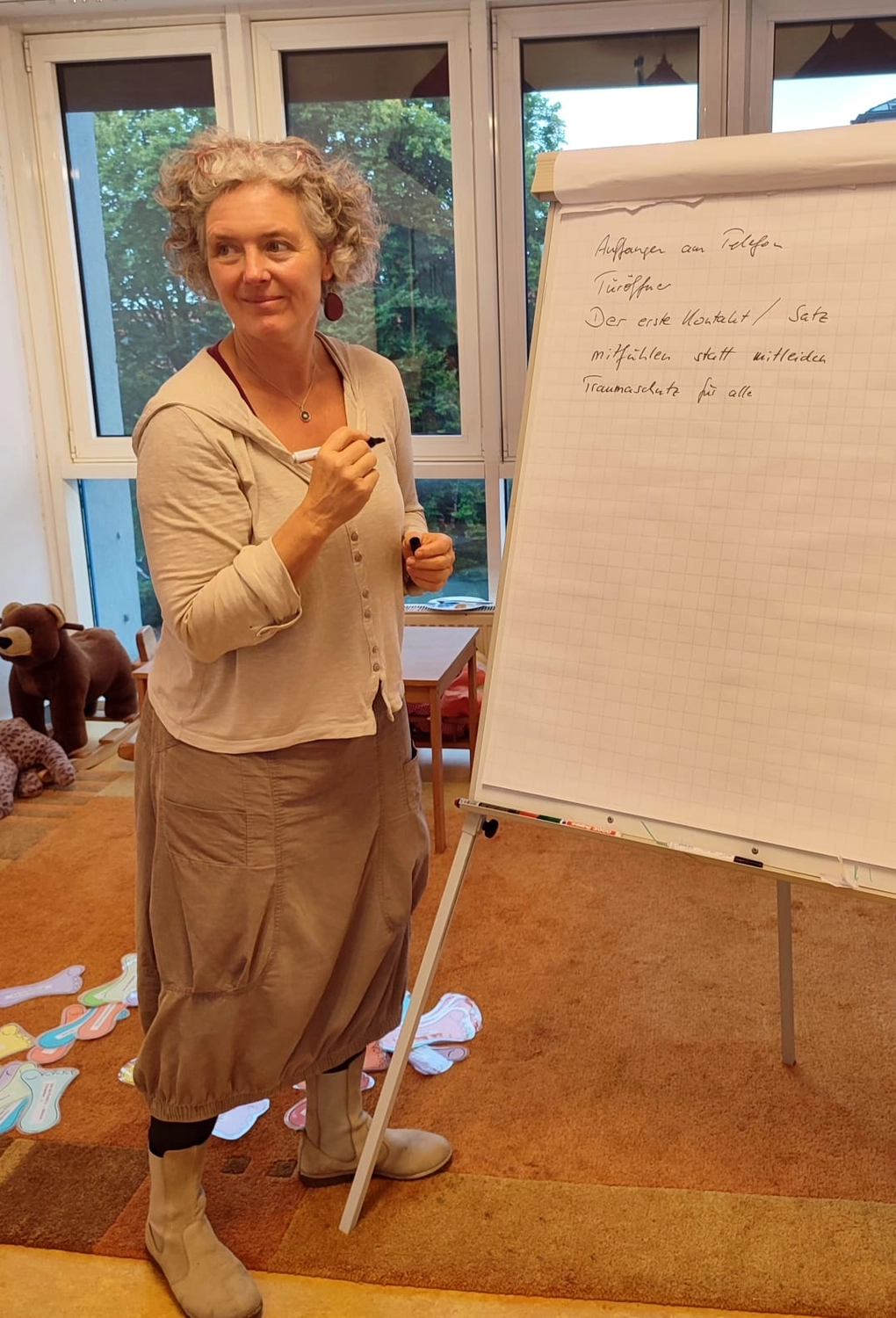Hebamme Trauma-Fachberaterin - Coaching, Beratung, Kurse und Lesungen - Eine Frau mit kurzen, lockigen grauen Haaren steht vor einer Tafel und schreibt darauf, während sie eine kleine Gruppe anspricht. Sie trägt eine dunkel gemusterte Jacke und einen langen, hautfarbenen Rock. Rechts hängt hinter einem leeren Stuhl ein farbenfrohes abstraktes Gemälde an der Wand.