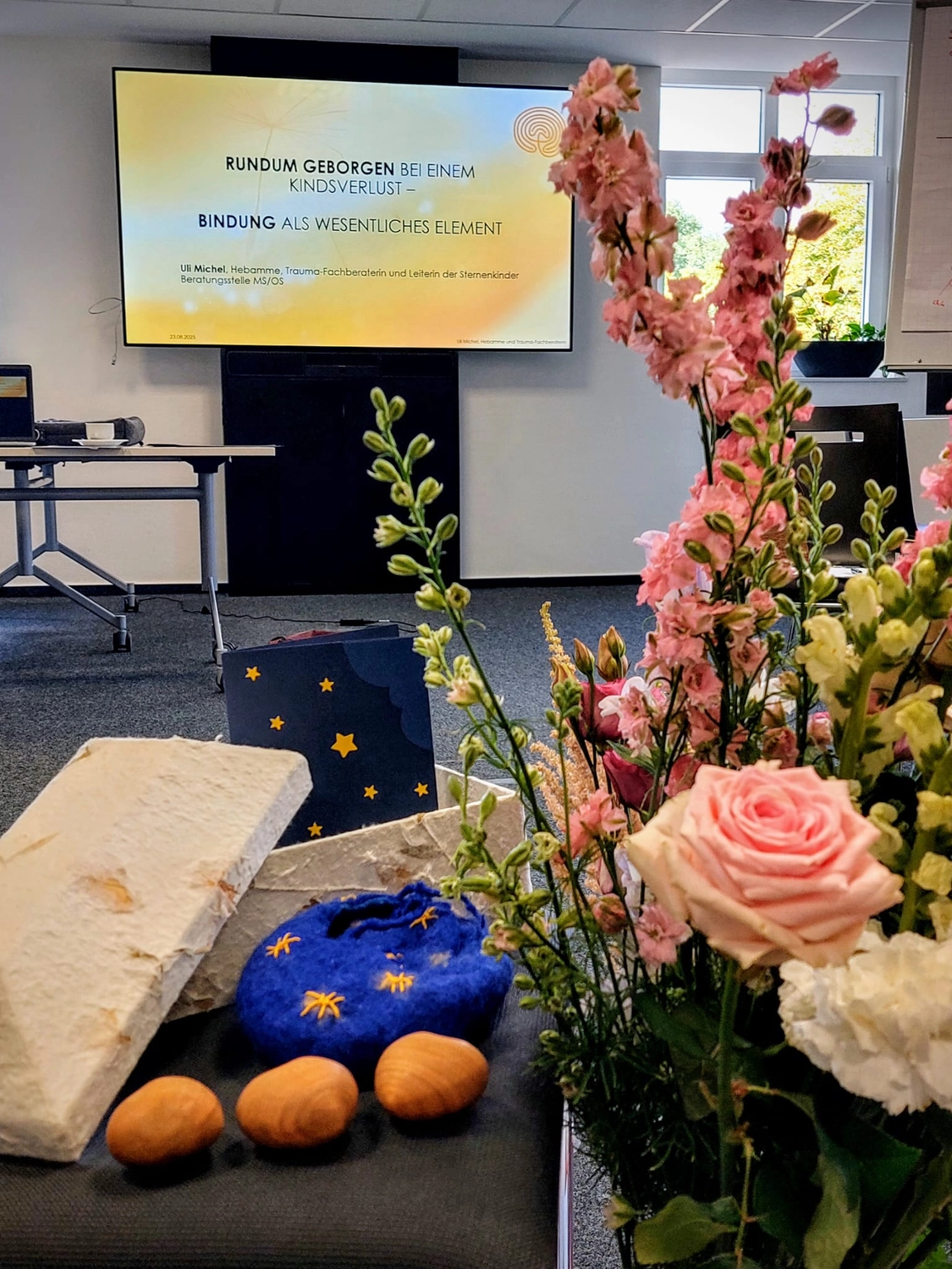 Hebamme Trauma-Fachberaterin - Coaching, Beratung, Kurse und Lesungen - In einem Raum mit Blumenarrangements, dekorativen Sternen und kleinen Holzeiern im Vordergrund ist ein Präsentationsbildschirm mit dem Titel "RUNDUM GEBORGEN BEI EINEM KINDSVERLUST" zu sehen.