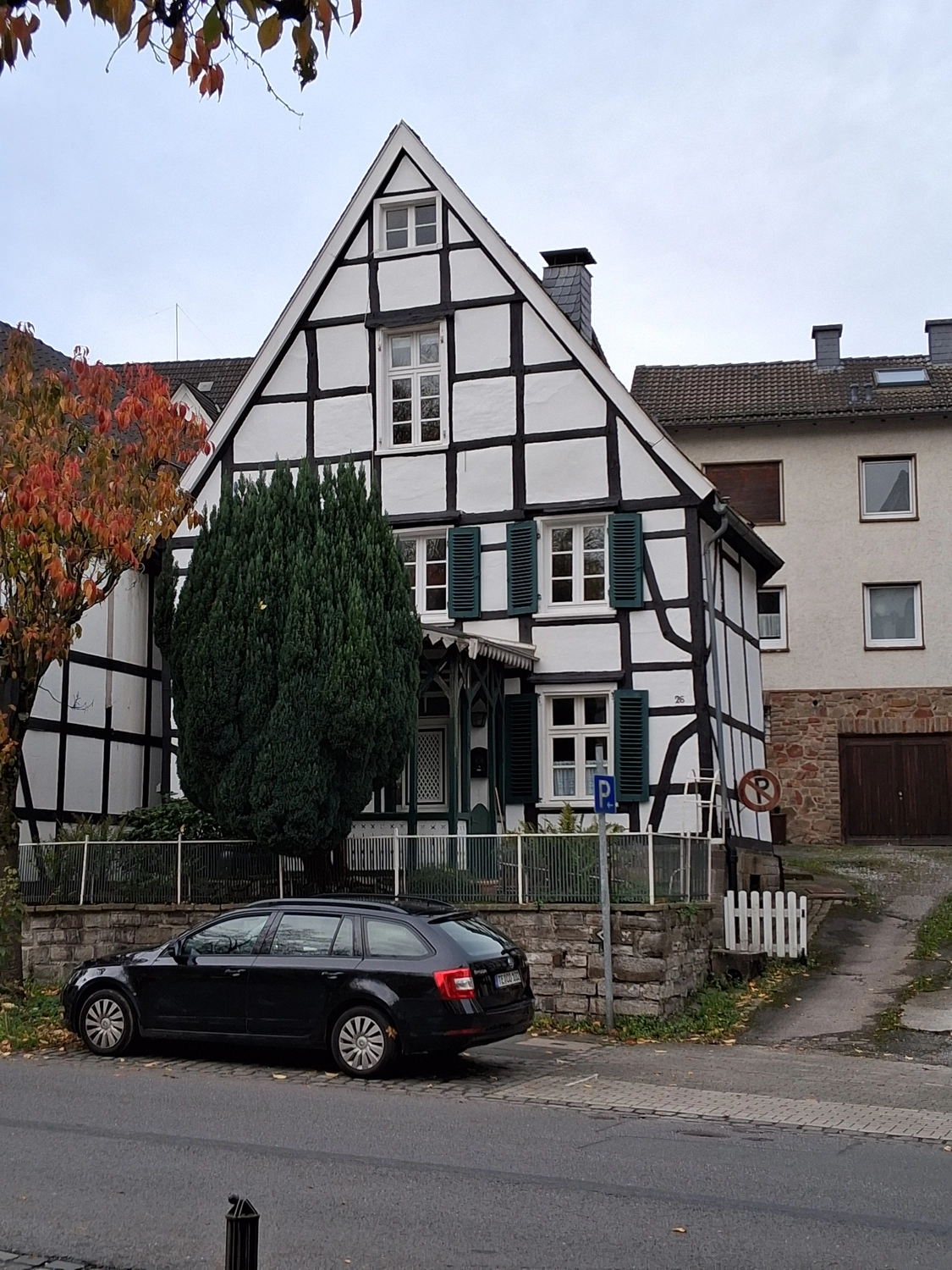 Hebamme Trauma-Fachberaterin - Coaching, Beratung, Kurse und Lesungen - Hinter einer Steinmauer und einem hohen immergrünen Baum steht ein schwarz-weißes Fachwerkhaus mit dunkelgrünen Fensterläden. Im Vordergrund parkt ein schwarzer Kombi auf der Straße.