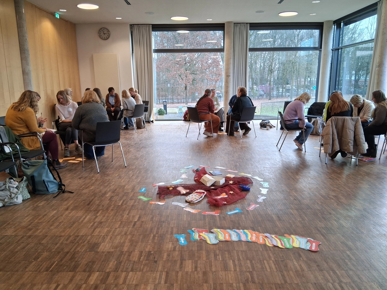Hebamme Trauma-Fachberaterin - Coaching, Beratung, Kurse und Lesungen - Ein großer, hell erleuchteter Seminarraum mit Holzboden und bodentiefen Fenstern, in dem sich eine große Gruppe von Menschen in kleinen Diskussionsrunden versammelt.