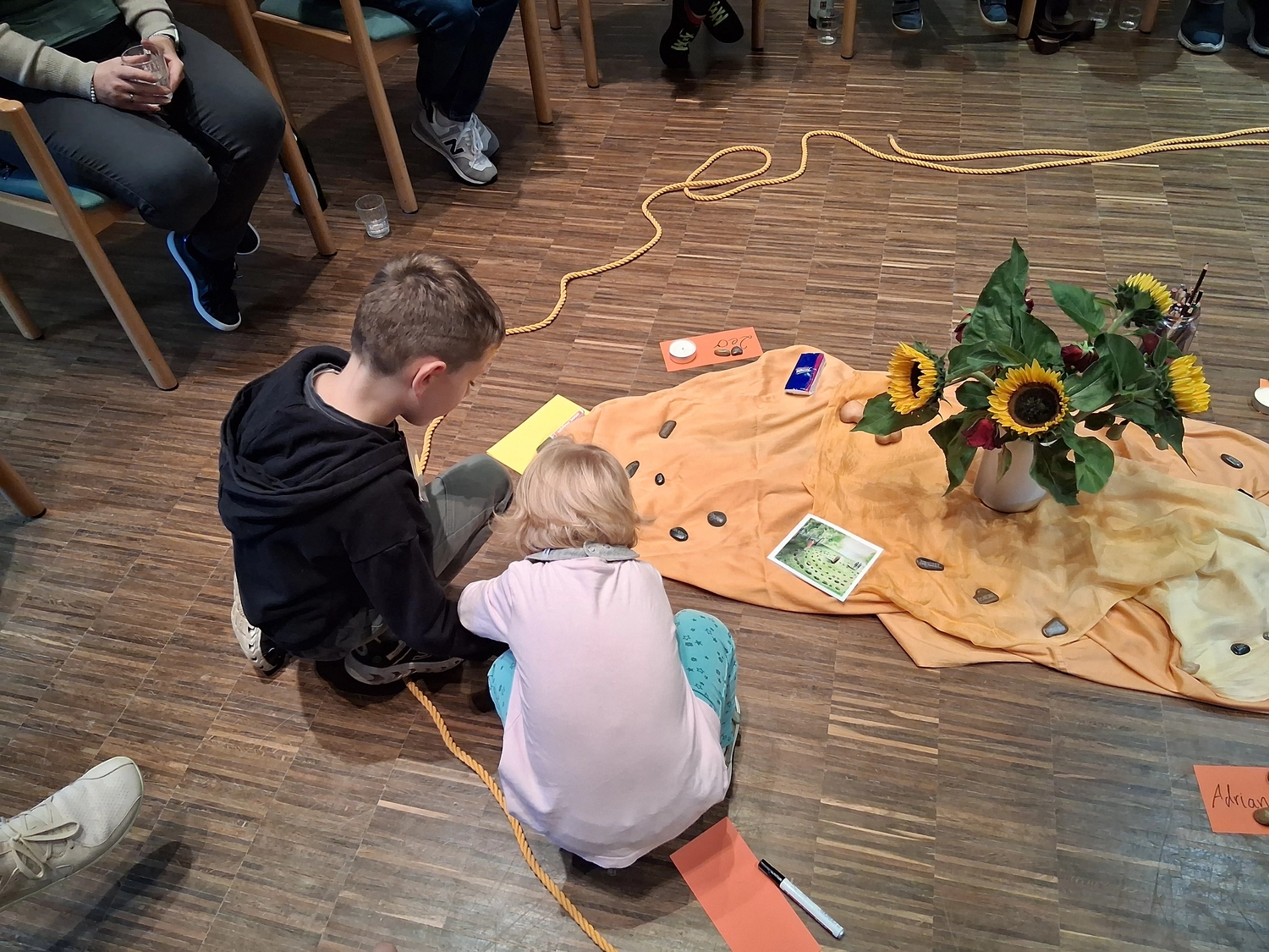 Hebamme Trauma-Fachberaterin - Coaching, Beratung, Kurse und Lesungen - Zwei Kinder sitzen auf einem Holzboden und betrachten konzentriert eine Anordnung von Steinen und Karten auf einem orangefarbenen Tuch, während eine Vase mit Sonnenblumen und ein aufgerolltes Seil die Szene einrahmen.