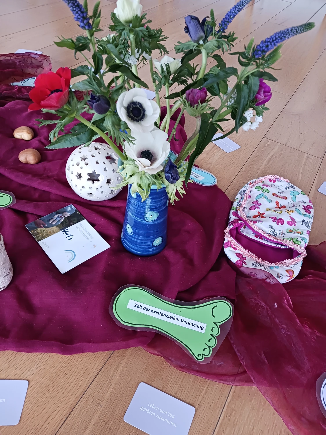 Hebamme Trauma-Fachberaterin - Coaching, Beratung, Kurse und Lesungen - Eine Aufnahme von oben zeigt ein Blumenarrangement in einer blauen Vase, umgeben von einer weißen Dekokugel, bunten Stoffen und bedruckten Karten auf einem Holzboden.