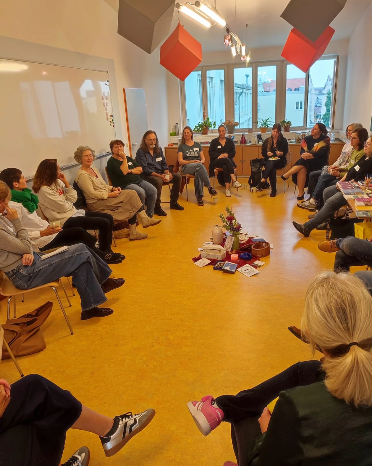 Hebamme Trauma-Fachberaterin - Coaching, Beratung, Kurse und Lesungen - Ein großer, hell erleuchteter Seminarraum mit Holzboden und bodentiefen Fenstern, in dem sich eine große Gruppe von Menschen in kleinen Diskussionsrunden versammelt.