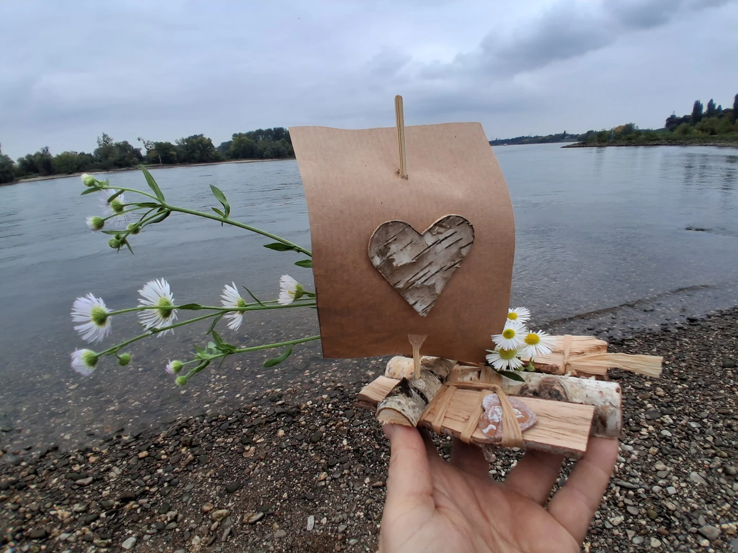 Hebamme Trauma-Fachberaterin - Coaching, Beratung, Kurse und Lesungen - Eine Hand hält ein kleines, handgefertigtes Spielzeugsegelboot aus Birkenholz und braunem Papier, verziert mit winzigen weißen Blümchen, vor dem Hintergrund eines Flusses unter einem bewölkten Himmel.