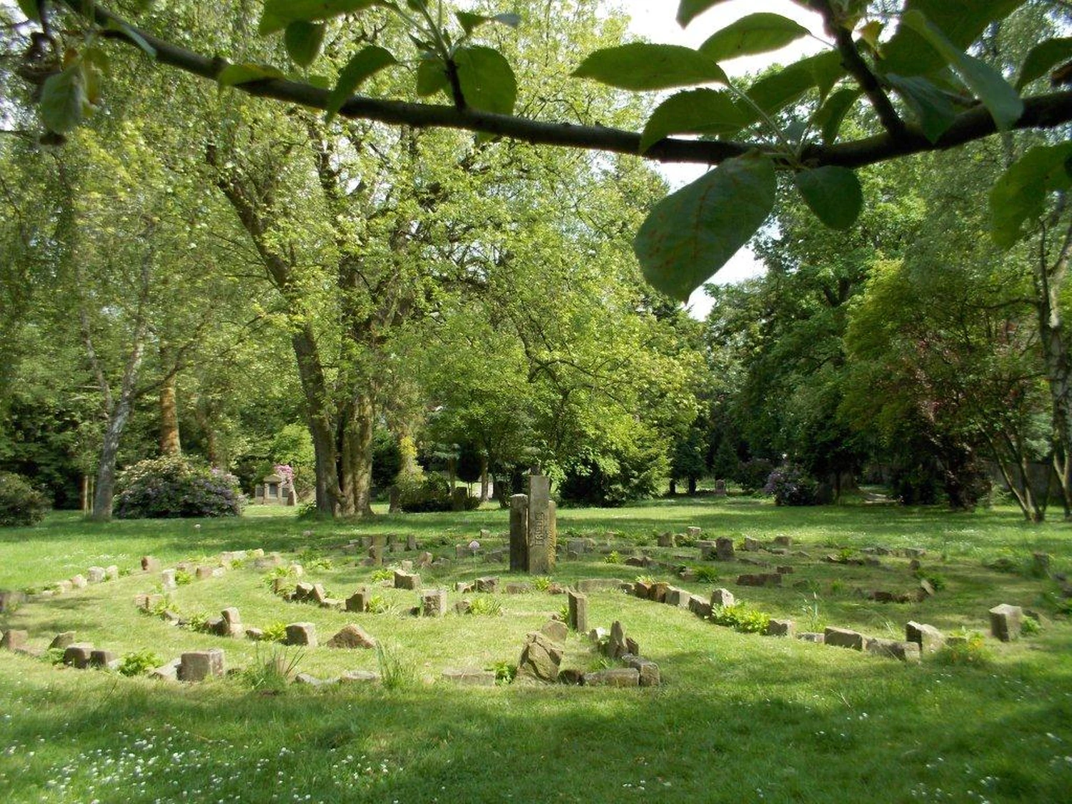 Hebamme Trauma-Fachberaterin - Coaching, Beratung, Kurse und Lesungen - Ein kreisförmiges Steinlabyrinth in einer üppigen, grünen Parkanlage. Das Labyrinth besteht aus niedrigen, verwitterten Steinen, die in konzentrischen Kreisen auf einer Rasenfläche angeordnet sind.