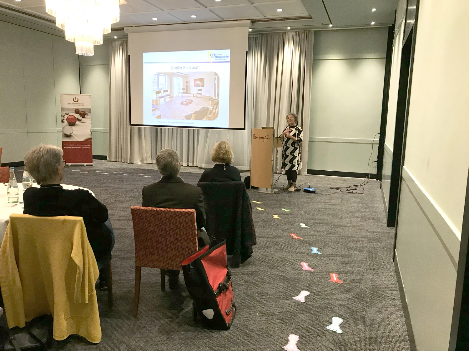Hebamme Trauma-Fachberaterin - Coaching, Beratung, Kurse und Lesungen - Eine Frau hält einen Vortrag vor einem kleinen Publikum in einem Konferenzraum mit einer Projektionsfläche und fußförmigen Aufklebern auf dem Boden.