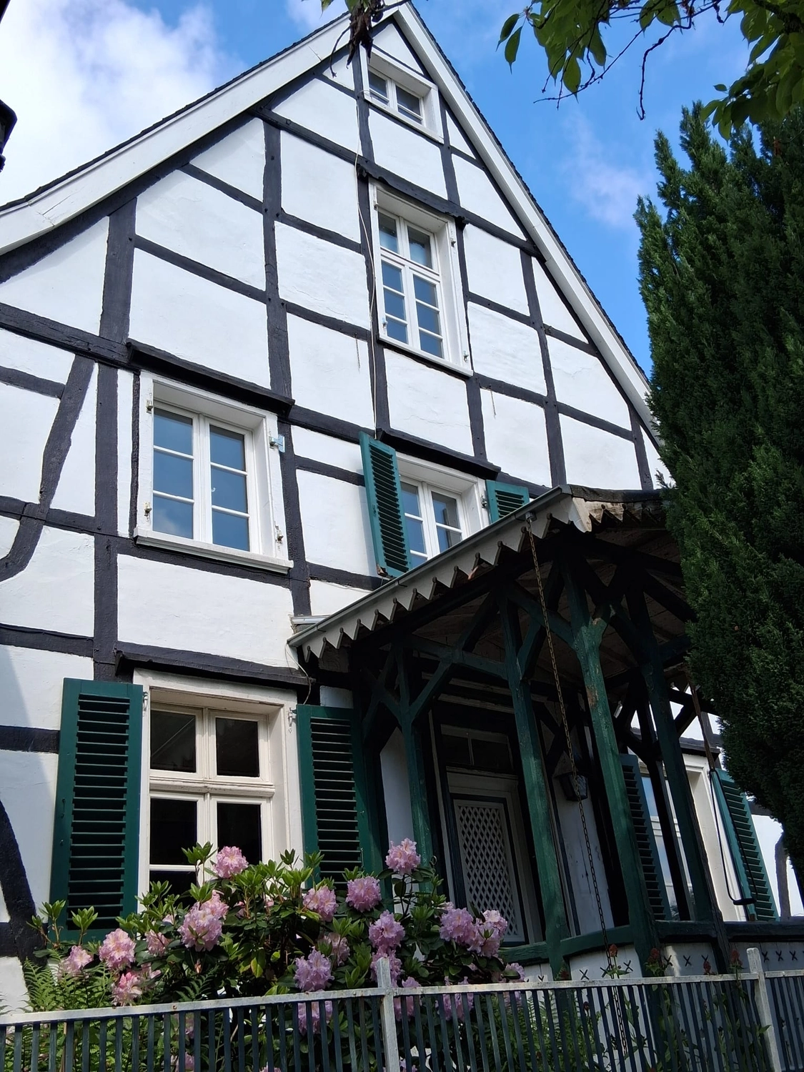 Aufnahme aus der Froschperspektive eines traditionellen weißen Fachwerkhauses mit dunklen Holzbalken unter einem strahlend blauen Himmel mit zarten Wolken.