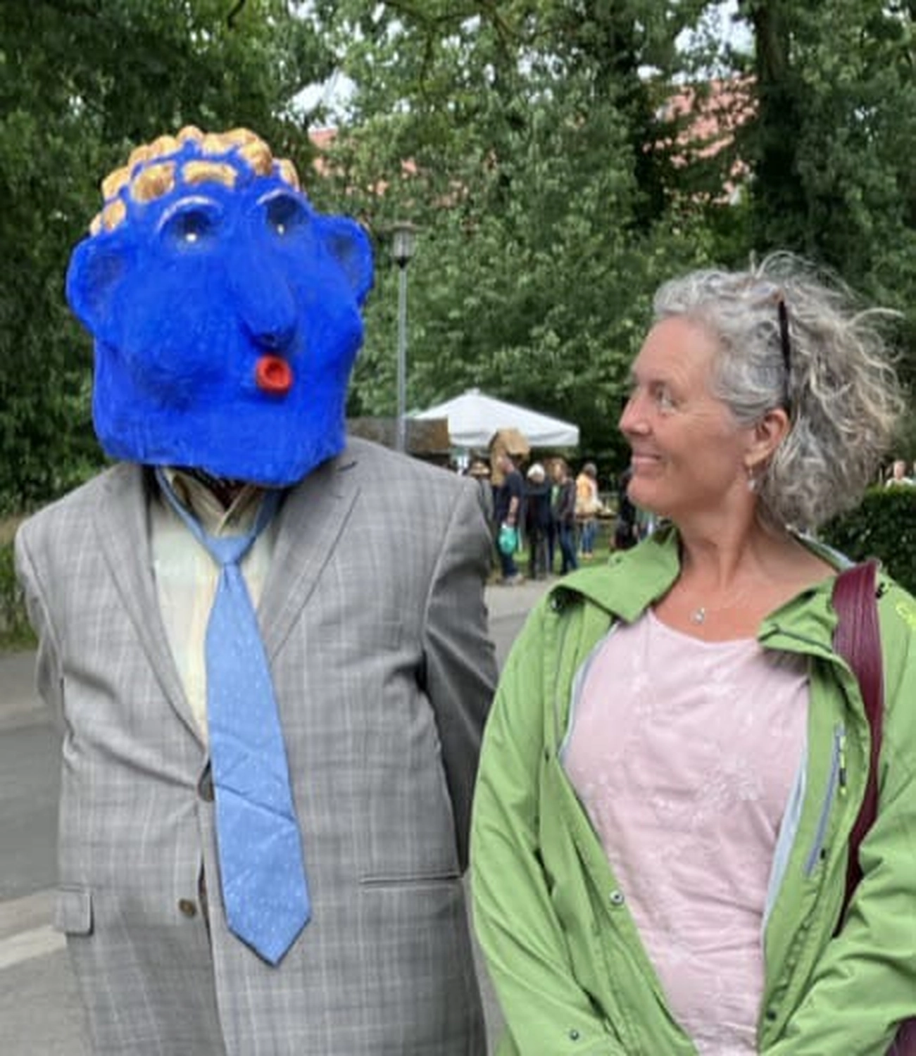 Hebamme Trauma-Fachberaterin - Coaching, Beratung, Kurse und Lesungen - Eine ältere Frau mit lockigem grauem Haar lächelt eine Person an, die eine große, strukturierte blaue Maske mit goldenem Oberteil und einen grauen Anzug mit blauer Krawatte trägt.
